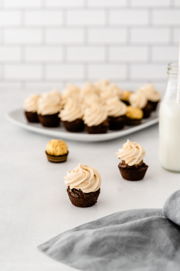 frosted ferrero rocher brownie bite with platter of brownies in the background and jar of milk