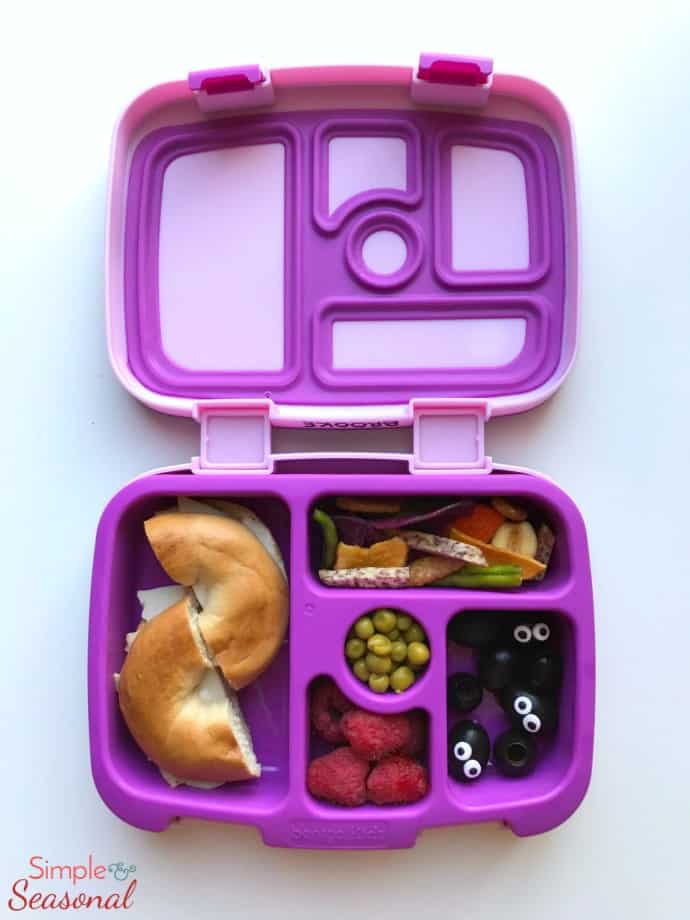 vertical image: bagel sandwich, raspberries, olives and dried veggie snacks in purple lunchbox