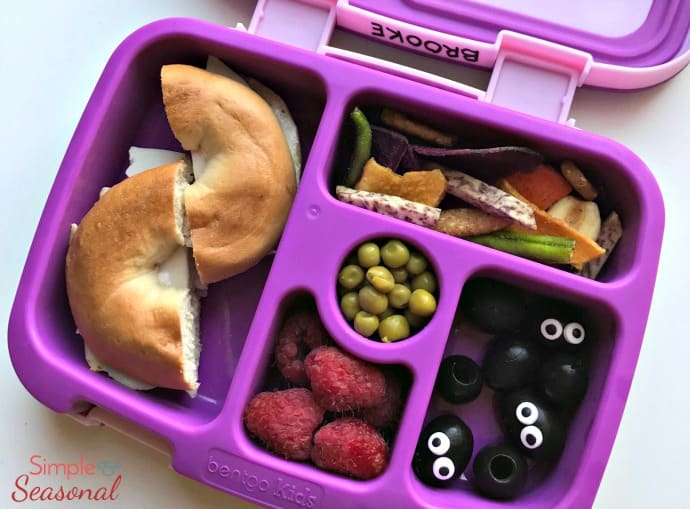 bagel sandwich, raspberries, olives and dried veggie snacks in purple lunchbox