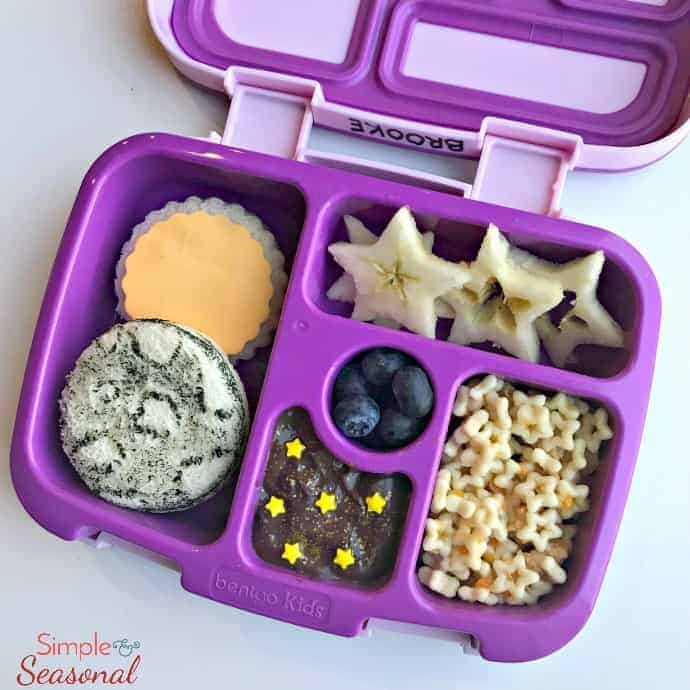 close up: sandwich decorated like sun and moon, star puffs, apple slices and other snacks in lunchbox 