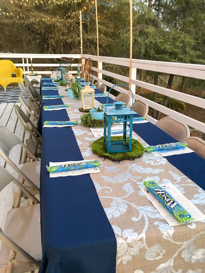 outdoor table with blue tablecloth, neutral runner, lanterns and green moss centerpieces