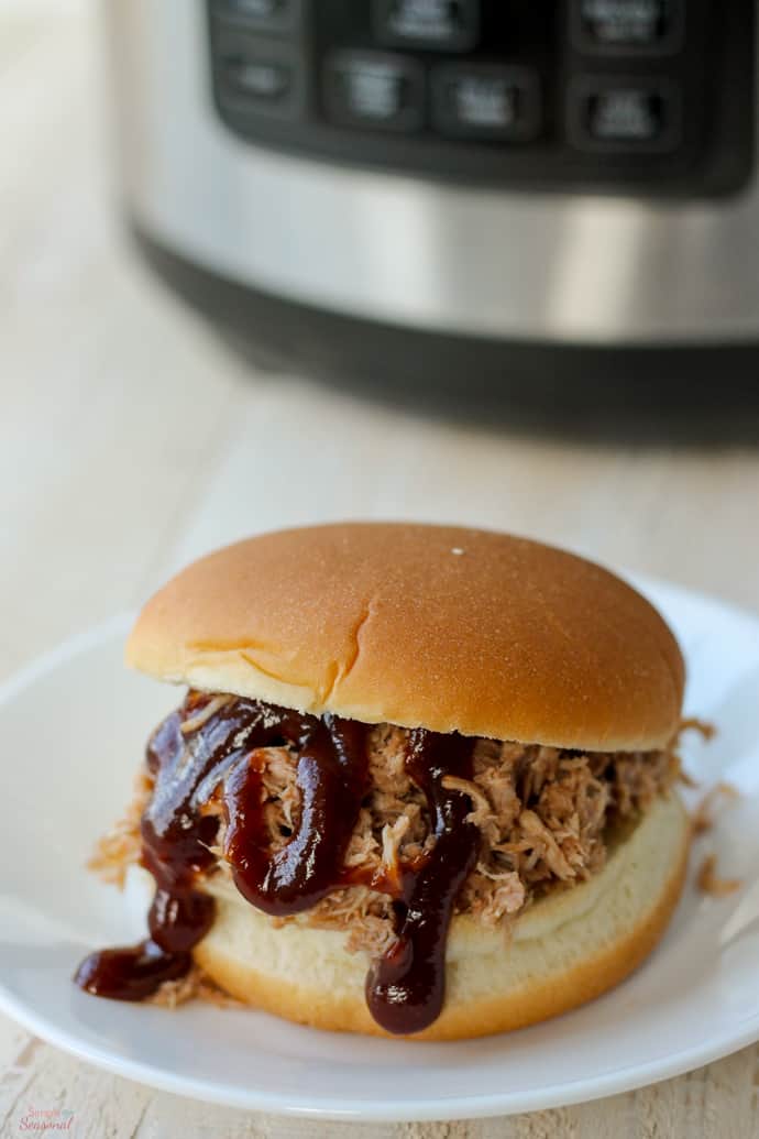 pulled pork sandwich on white plate in front of pressure cooker