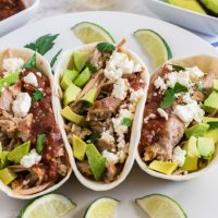 close up shot of taco shells holding pork and other toppings with lime wedges as garnish