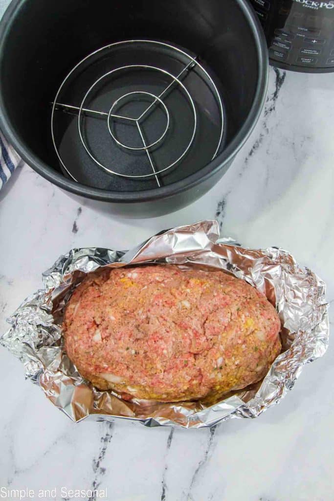 raw meatloaf wrapped in foil, pressure cooker pot with trivet inside and water on the bottom