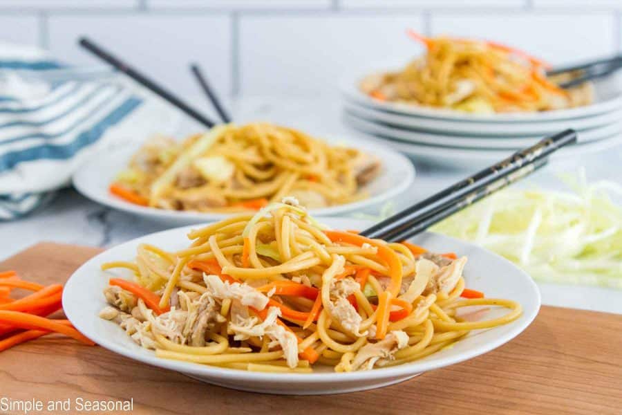 plate of noodles, chicken and vegetables with stack of plates in the background