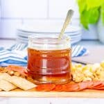 jar of honey with a spoon on a board with snacks; stack of plates in background