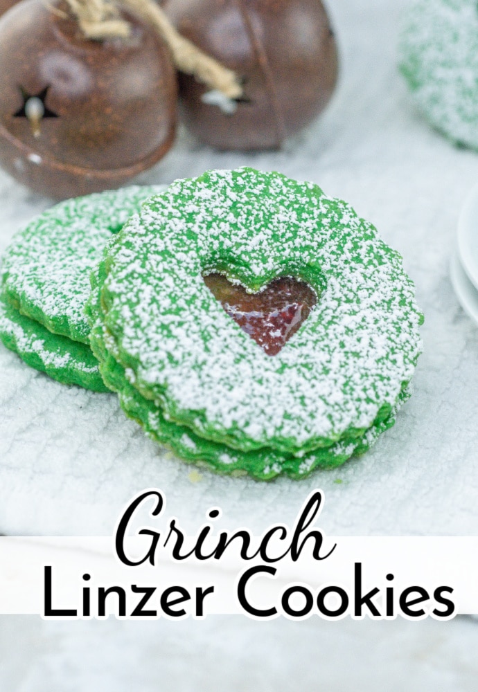 green Grinch heart cookie leaning up against another cookie with Christmas bells in the background; text reads Grinch Linzer Cookies