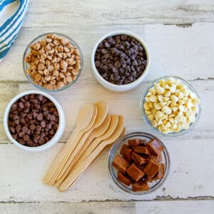 bowls of baking chips and caramels alongside a stack of flat wooden spoons-ingredients for hot cocoa spoons