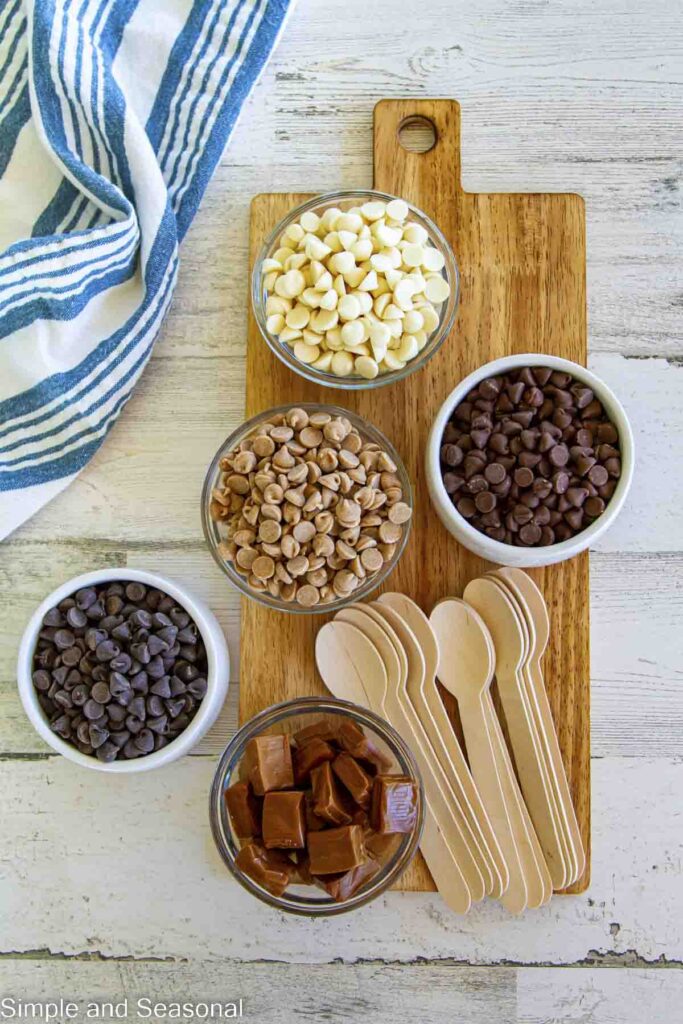 variety of baking chips and caramels for melting, along with wooden spoons on a platter