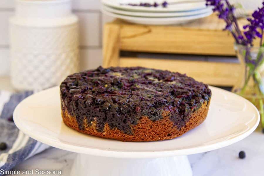 whole round cake on a white platter with purple flowers in the background