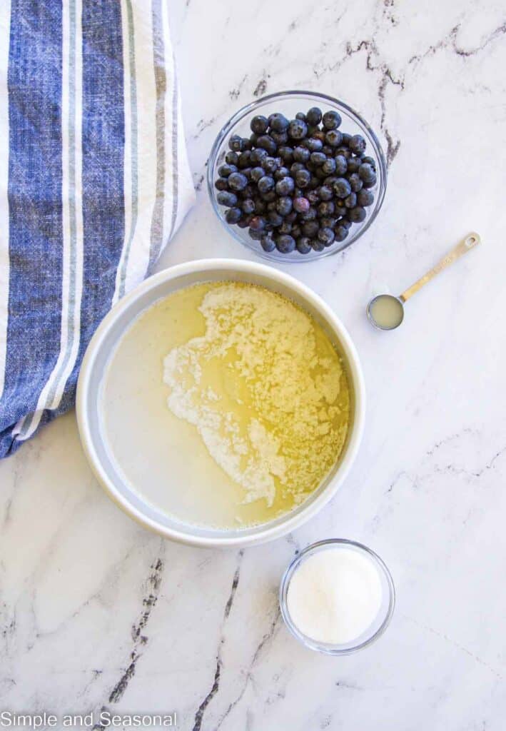 round cake pan with melted butter inside and blueberries, sugar and lemon juice ready to add